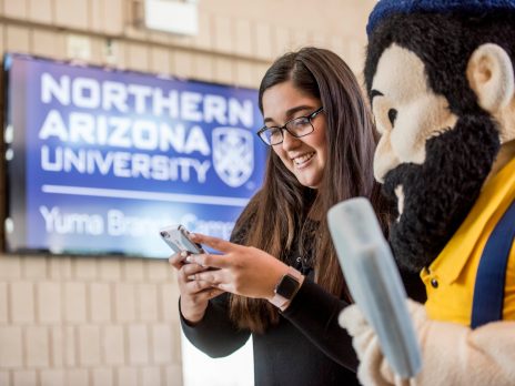 NAU student on her phone standing next to the Louie the Lumberjack mascot with a NAU sign in the background
