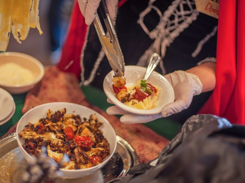Torso of person in cultural clothing serving what appears to be a cultural dish of some sort, in a bowl using tong. Pasta, meat and vegetables are featured in the dish.