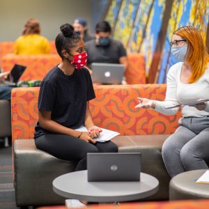 Two students talk in a common space while wearing masks