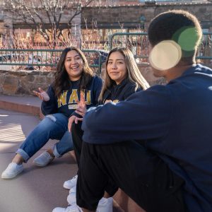 Three students sit outside in downtown Flagstaff, wearing NAU sweatshirts