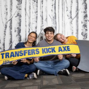 Three students sit in the hallway with a sign that reads "Transfers kick axe"