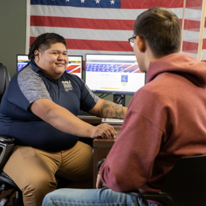 A student sits at a desk with a VMS staff member, in front of an American flag