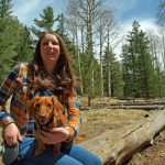 Photo of Kimberly Walker sitting with a dog on her lap. There are pine trees in the background.