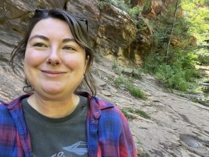 Photo of Mirra wearing a grey t-shirt and blue and red jacket standing in front of a rock outcropping