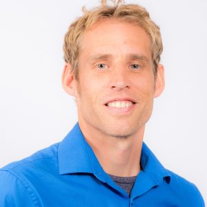 Professional headshot of Dr. Dirk de Heer wearing a bright blue button up