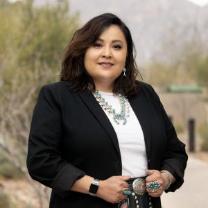 Monica Yellowhair wearing a printed skirt, white top, and black overcoat with a flourish of turquoise and silver jewelry