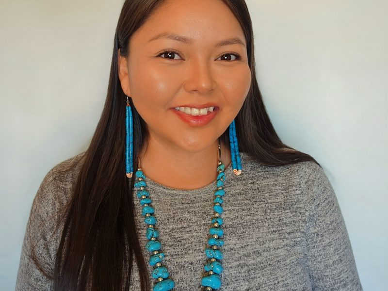 Joyceline Greymountain wearing a long sleeve grey top with a large turquoise necklace and long turquoise earrings.