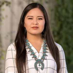 Headshot photo of the Dynnika Tso wearing a white and black long sleeve top and turquoise squash blossom with matching earrings