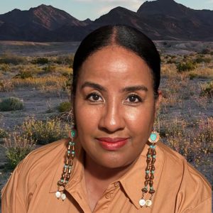 Headshot photo of Darnella Melancon wearing a tan top with copper and turquoise earrings and a southwest landscape background