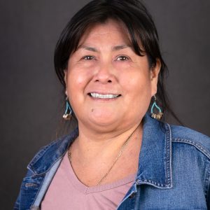 Headshot photo of Inez Zabala wearing a pink top and jean jacket with turquoise earrings and a dark gray background.