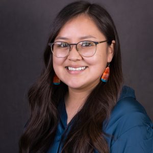 Headshot photo of Jennifer Hudson wearing a dark teal top, coral earrings, and glasses.