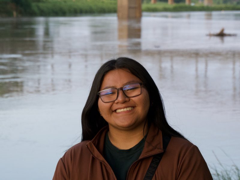 Headshot photo of Autumn Russell wearing a brown long sleeve top and dark brown glasses with a bridge and river in the background