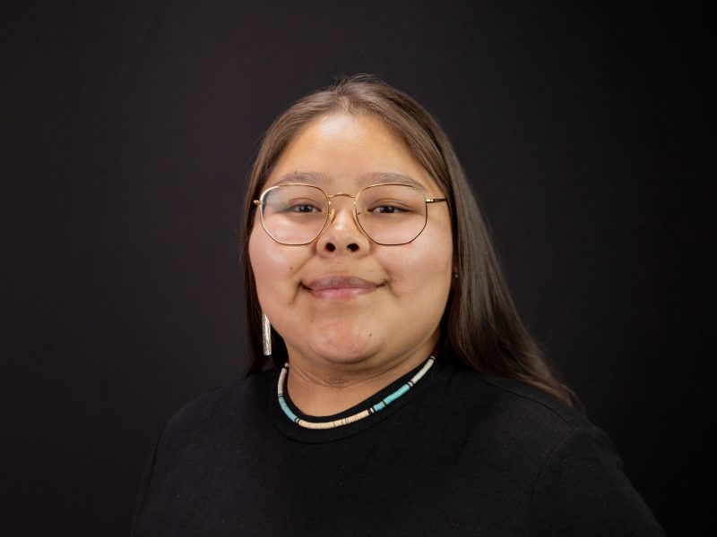 Headshot photo of Lortis Eriacho wearing gold glasses with a black top, turquoise necklace and silver earrings.