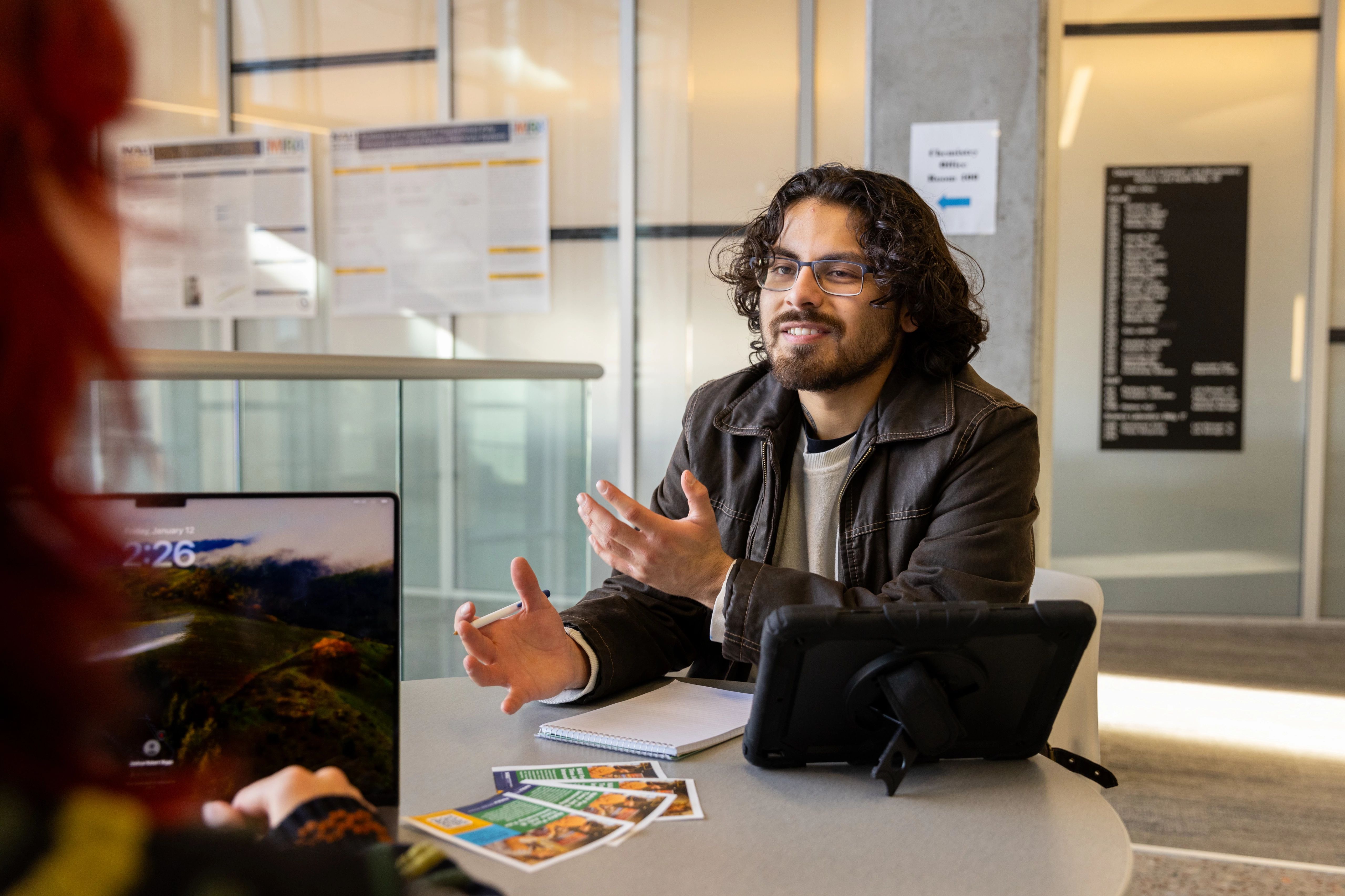 A male student sitting at a table working on papers and a laptop while talking to another student.