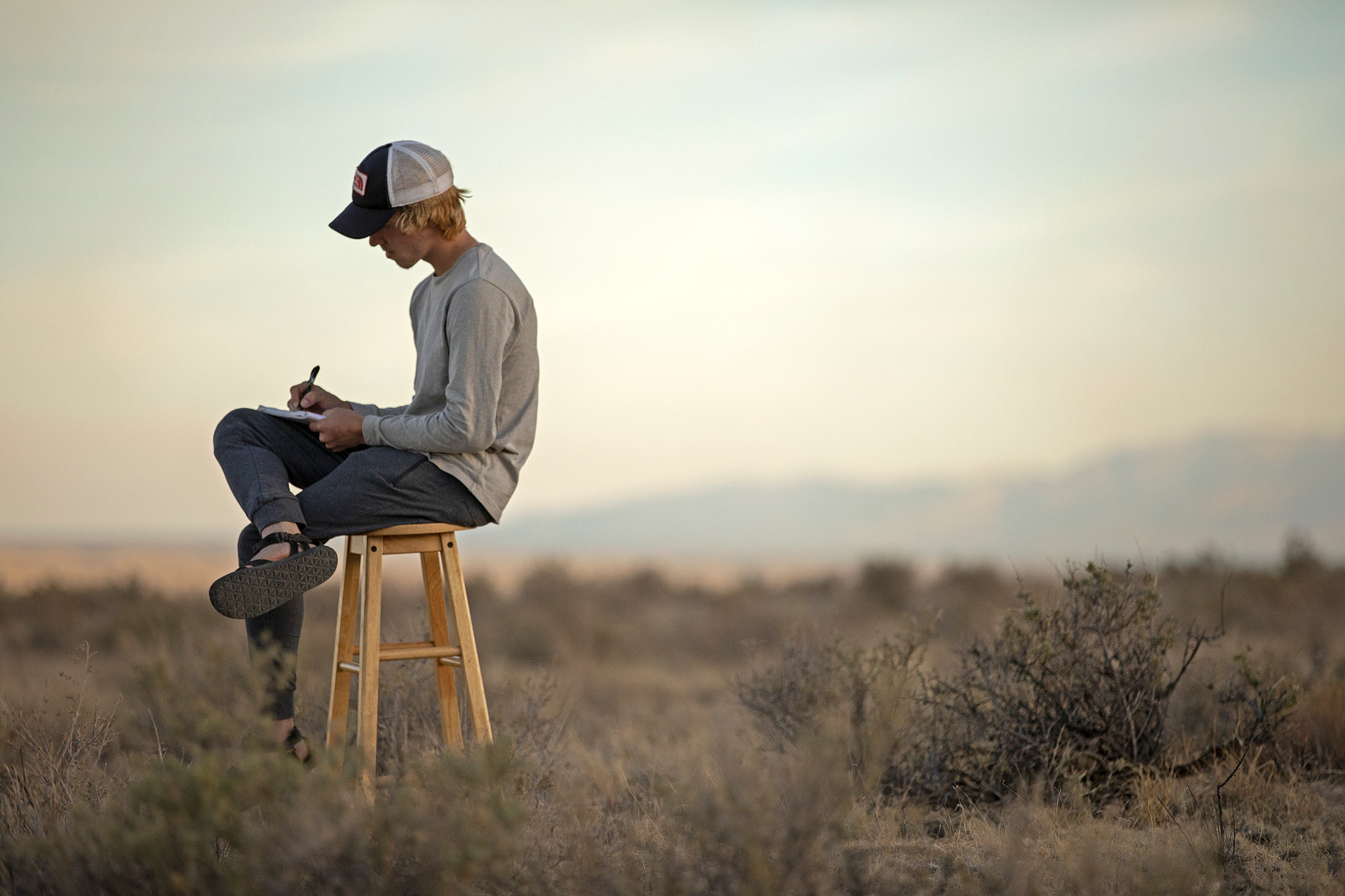 Student writes in a journal while sitting on a stool in a field.