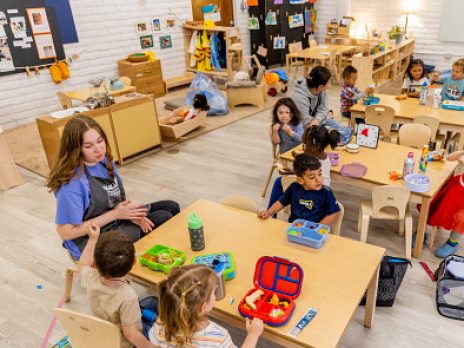 Several children and teachers eating lunch together at wooden classroom tables.