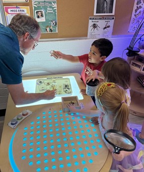 Three children and a professor at the light table. A child is holding up a bug specimen for the professor to see.