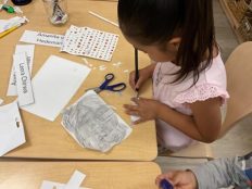 A child drawing with a colored pencil, with scissors, stickers, and name tags on the table surrounding her.