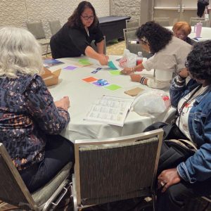 Four women sitting at a table playing with robot cars and color mats. One lady is pointing to the car to facilitate engagement from the others.