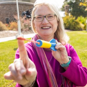 A female with glasses, Dr. Canipe, holding out a rubber band rocket, pretending to shoot it at the camera.