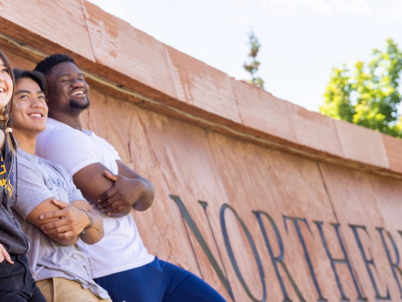 Five students smile and lean against the south Northern Arizona University sign.
