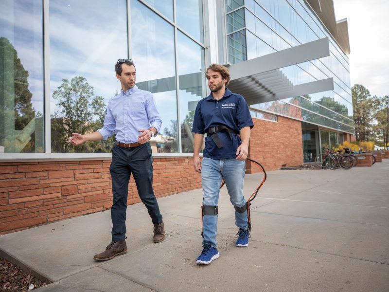 Two men walking, one with a walking aid machine strapped to him.