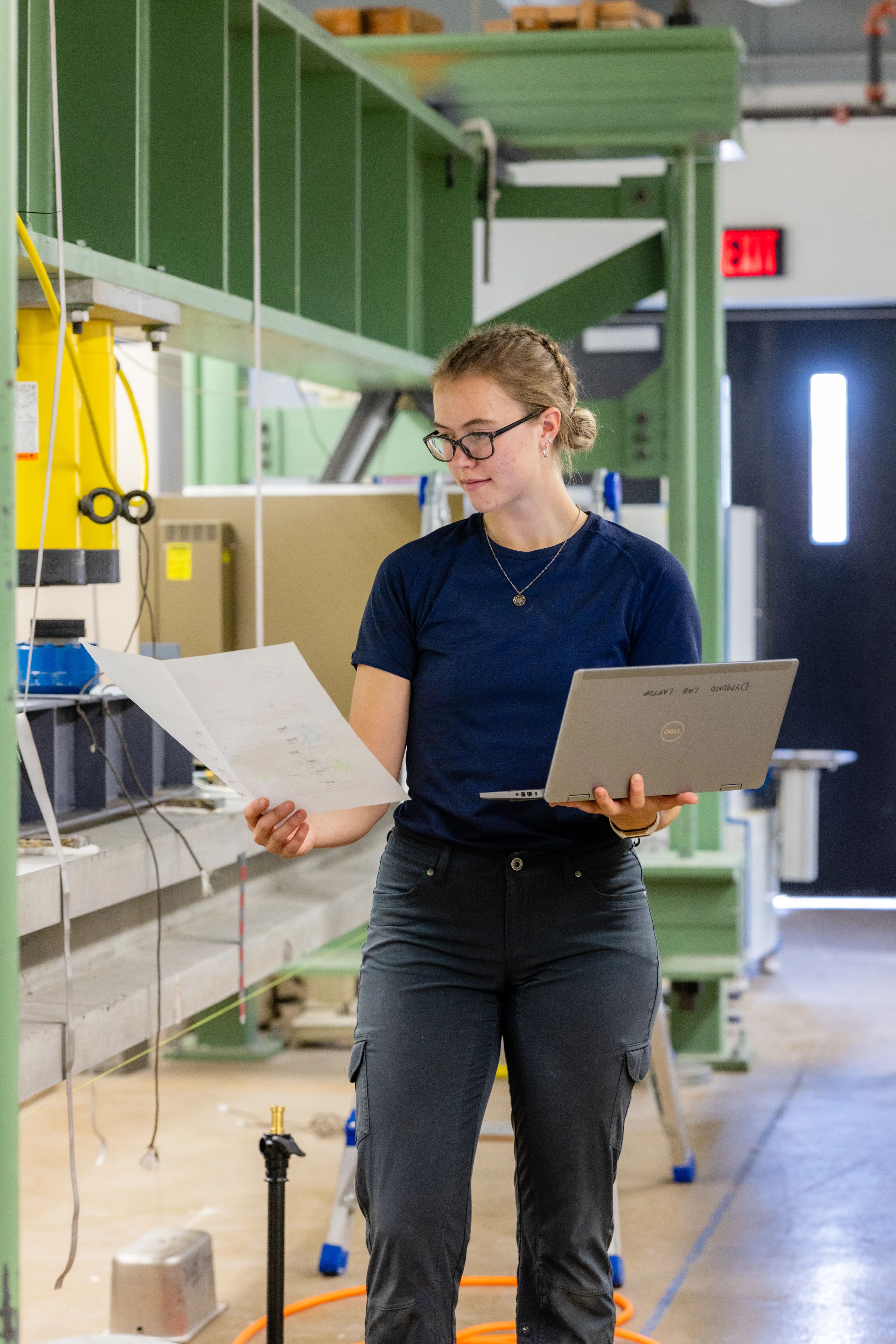 Student walking through a lab with a paper in one hand and a laptop in the other.