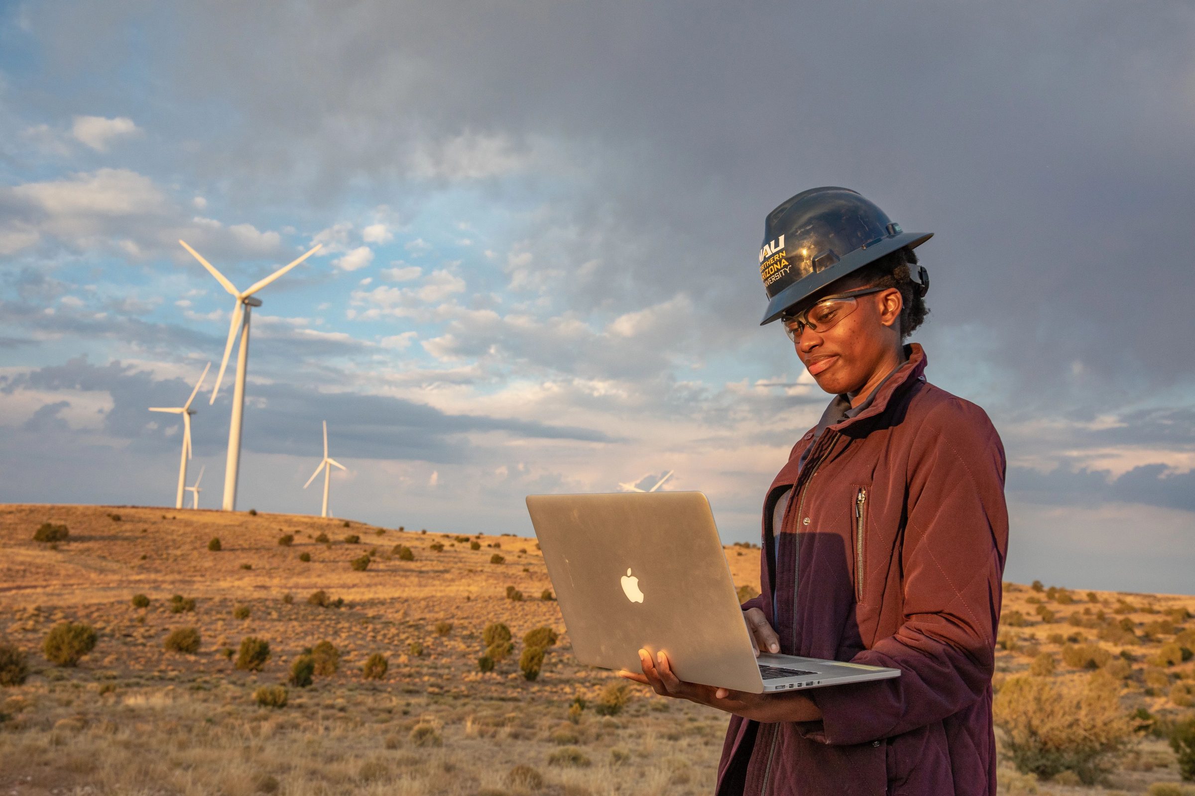N A U student standing outside at a Wind Farm and looking down at a laptop.