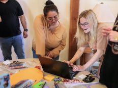 Two female students smile as they work together and lean over an open laptop. The table where the laptop sits is covered in colorful construction paper and pipecleaners.