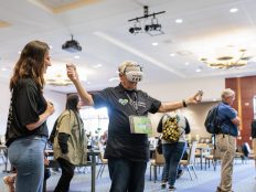 A researcher stands next to an older gentlemen wearing a virtual reality headset. His arms are outstretched to each side, holding controllers.