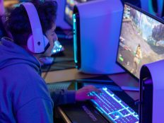 Person wearing headphones and sitting at a computer desk, playing a video game on a large screen monitor with colorful lighting. The person is focused on the game and has a keyboard and gaming mouse on the desk.