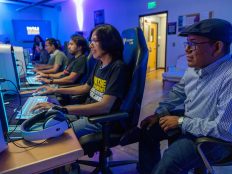 A group of people sitting at computers in a brightly lit room, focused on their screens. The room appears to be some kind of computer lab or gaming area, with monitors, keyboards, and gaming chairs visible. The people are wearing casual clothing and appear to be engaged in various activities on the computers.