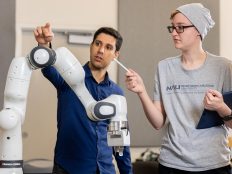 Two individuals wearing casual clothing, one in a blue shirt and the other in a grey shirt, examining a robotic arm.