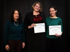 Three people standing together, two women and one person, holding certificates in their hands. All are wearing formal attire and the background is dark.