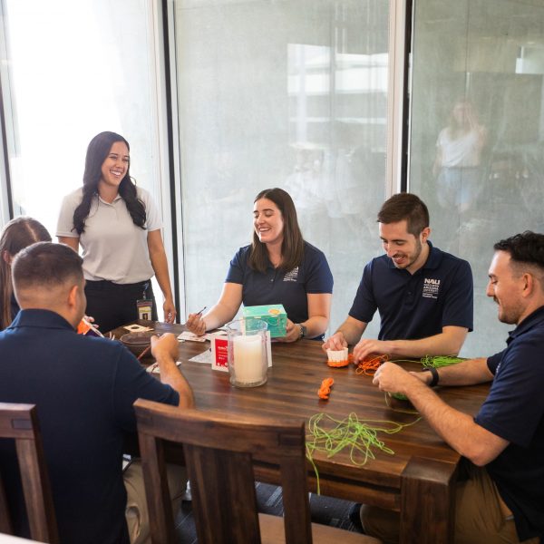 Click image to be directed to PBC Library services. A group of students sit around a dark wooden table smiling and conversing as they work on program material. 