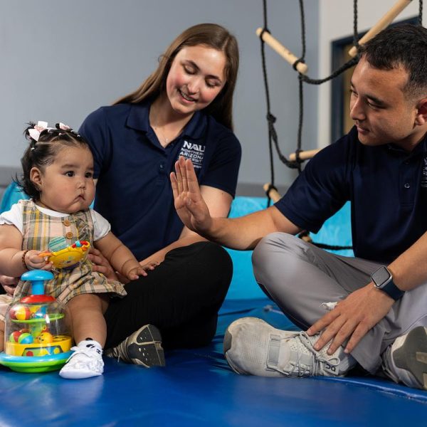 Click image for redirection to NAU scholarships. Young female toddler with toys an bows in her hair sits on the lap of a female occupational therapist while a male therapist sitting to the right high-fives her. 