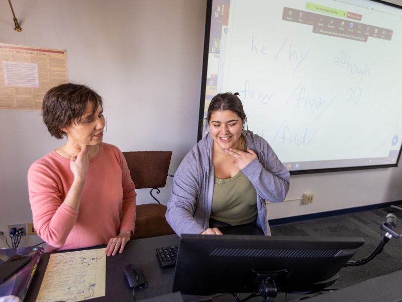 A professor and student practice pronunciations while holding two fingers to their neck.
