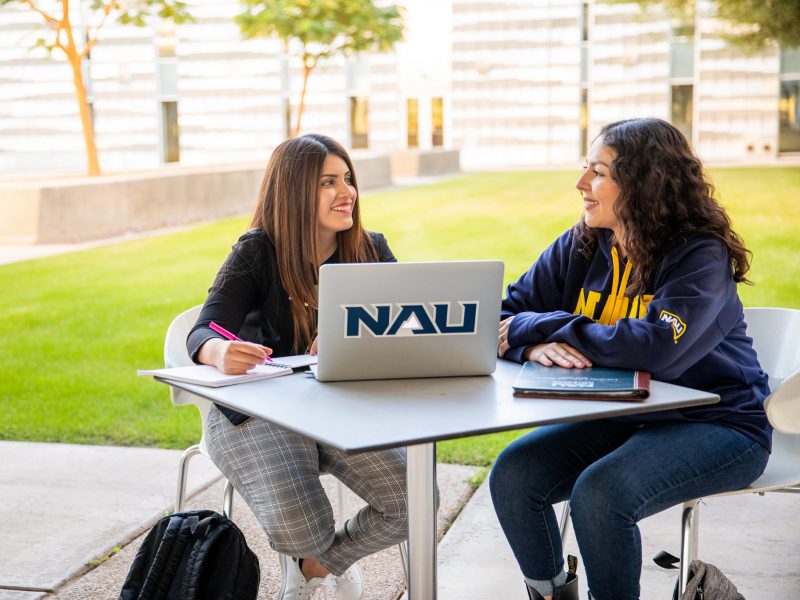 Two students sit at a table outside and work at a computer.