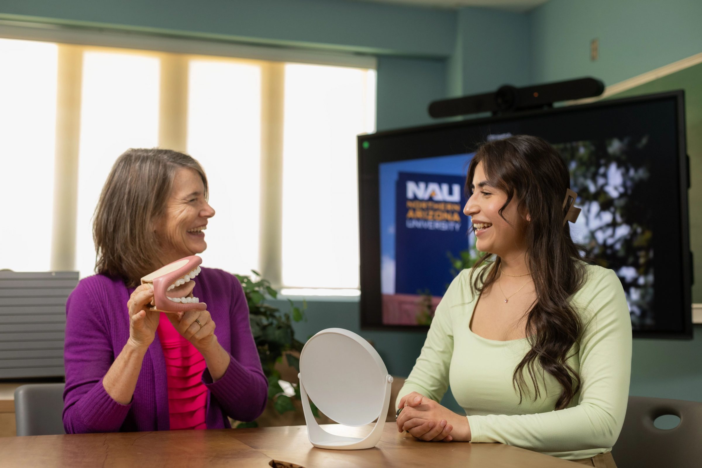 A professor and student sit at a table in front of a small mirror.