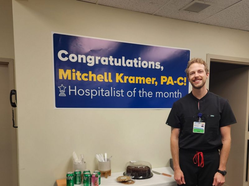 Mitchell Kramer, graduate of the N A U Physician Assistant Studies program, standing next to a congratulations banner for Hospitalist of the Month for MedStar Health.