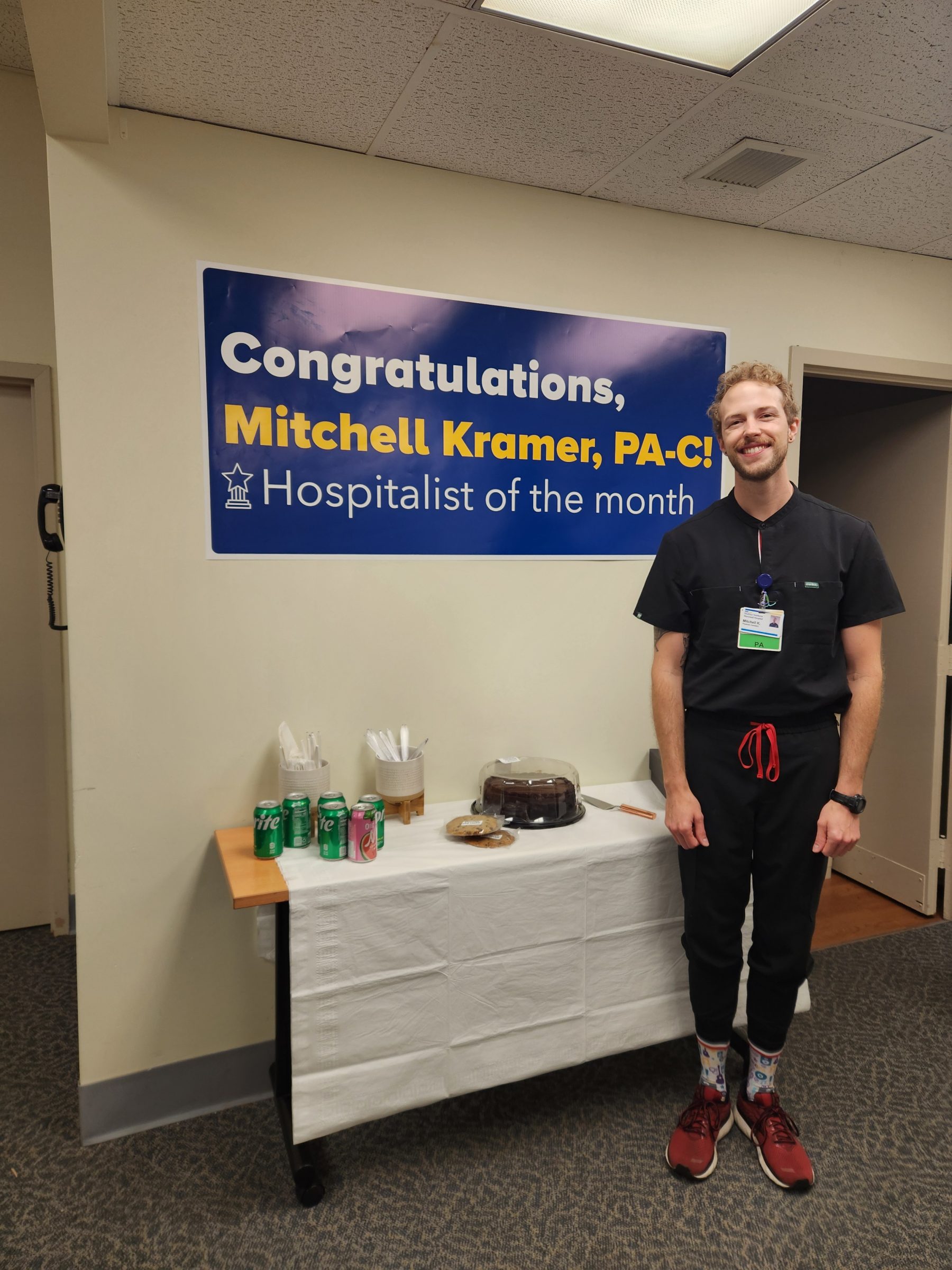 Mitchell Kramer, graduate of the N A U Physician Assistant Studies program, standing next to a congratulations banner for Hospitalist of the Month for MedStar Health.