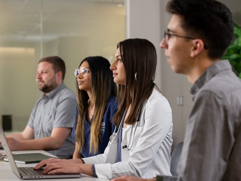 Four students and professionals sit side by side at a conference table, focused on a discussion or presentation. Two participants work on laptops, while one wears medical scrubs and another wears a white coat with a stethoscope, suggesting a healthcare-related learning or collaborative environment.