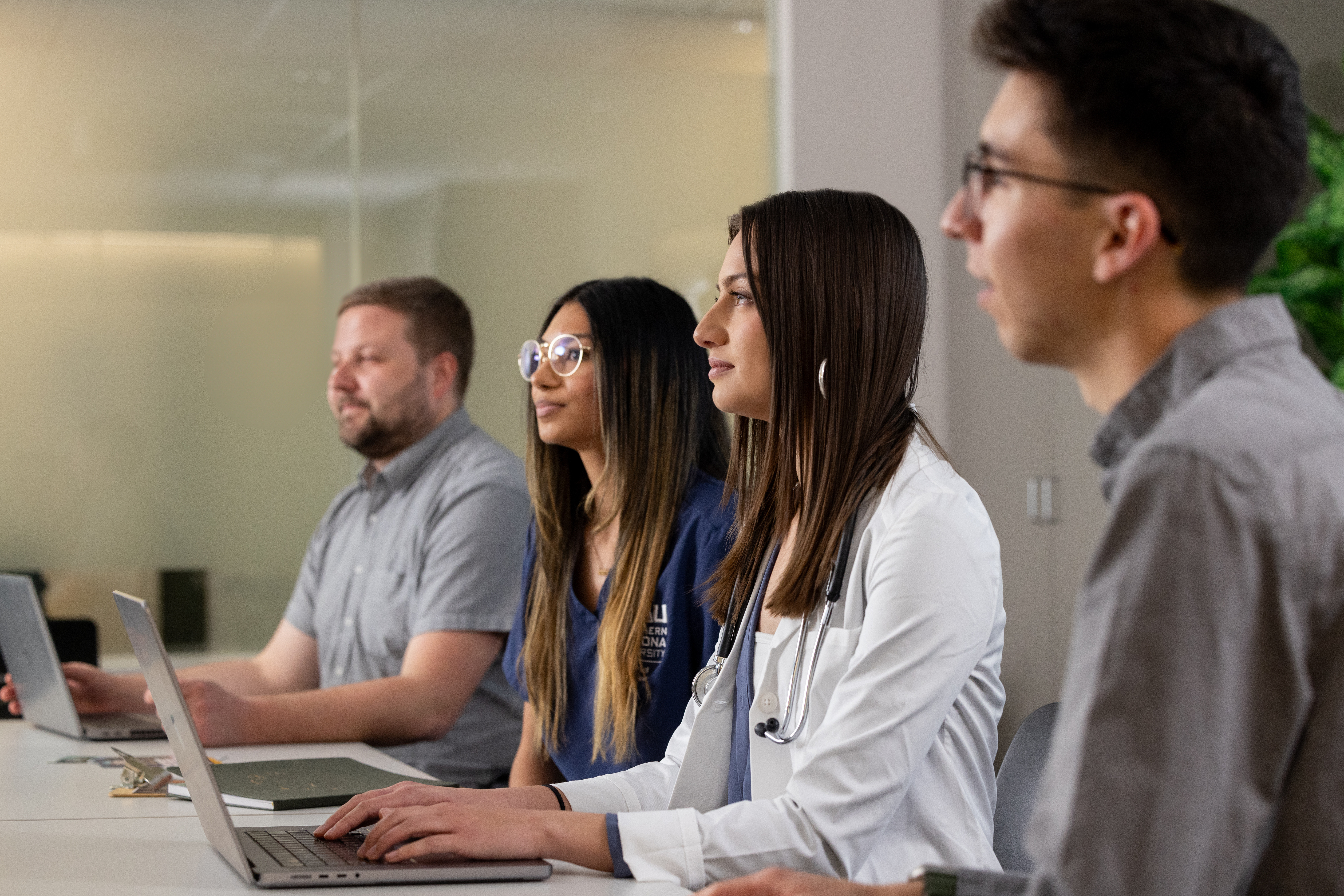 Four students and professionals sit side by side at a conference table, focused on a discussion or presentation. Two participants work on laptops, while one wears medical scrubs and another wears a white coat with a stethoscope, suggesting a healthcare-related learning or collaborative environment.