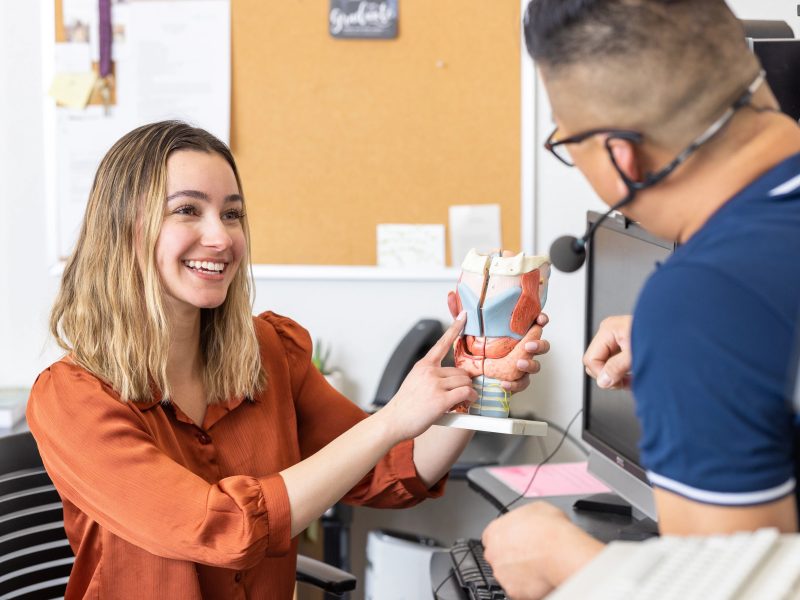 A speech-language pathologist smiles while holding an anatomical model of the throat and vocal tract, pointing to its structures as they explain them to a patient during a consultation in a clinical office. The patient sits across the desk wearing a headset microphone, listening attentively.
