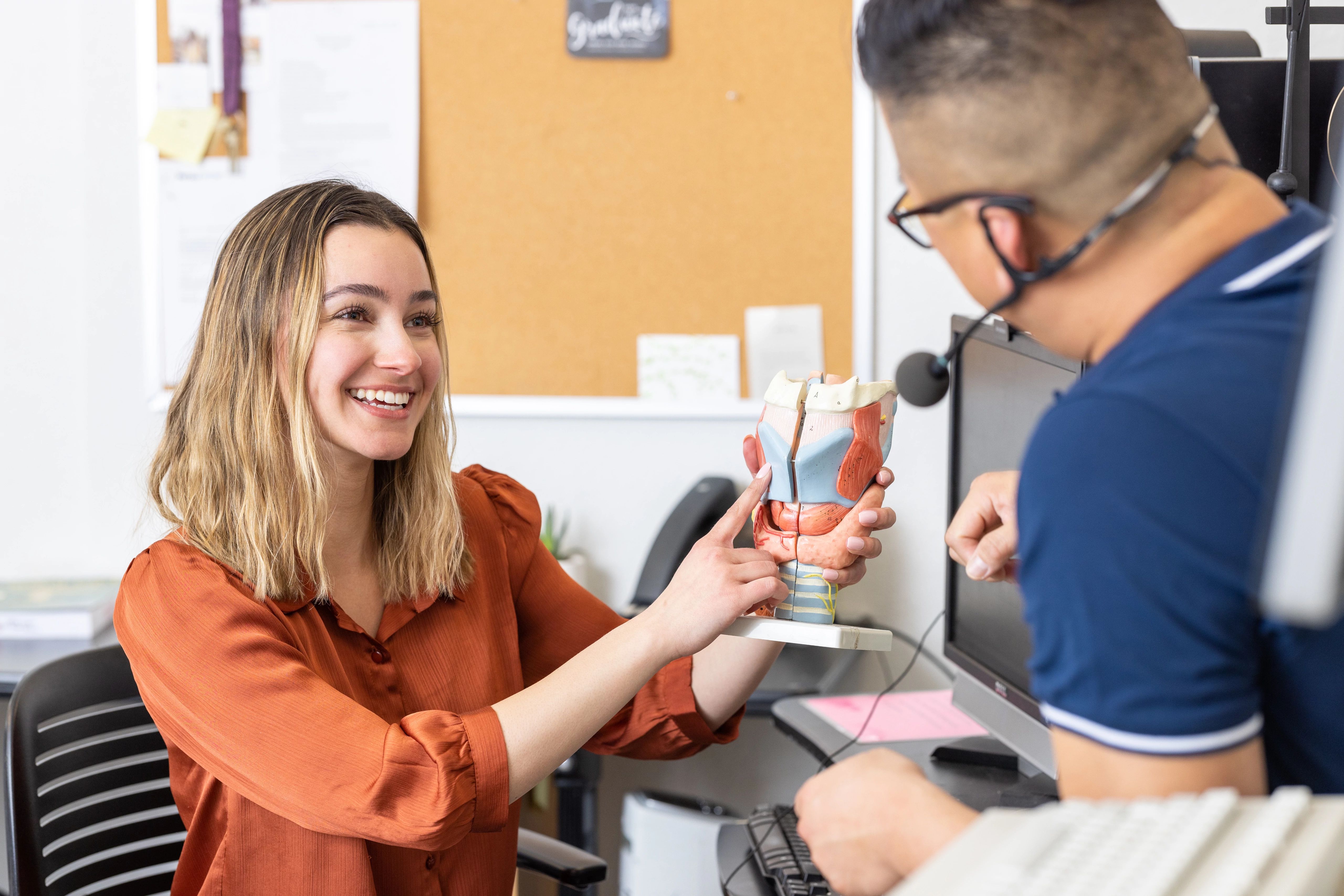 A speech-language pathologist smiles while holding an anatomical model of the throat and vocal tract, pointing to its structures as they explain them to a patient during a consultation in a clinical office. The patient sits across the desk wearing a headset microphone, listening attentively.
