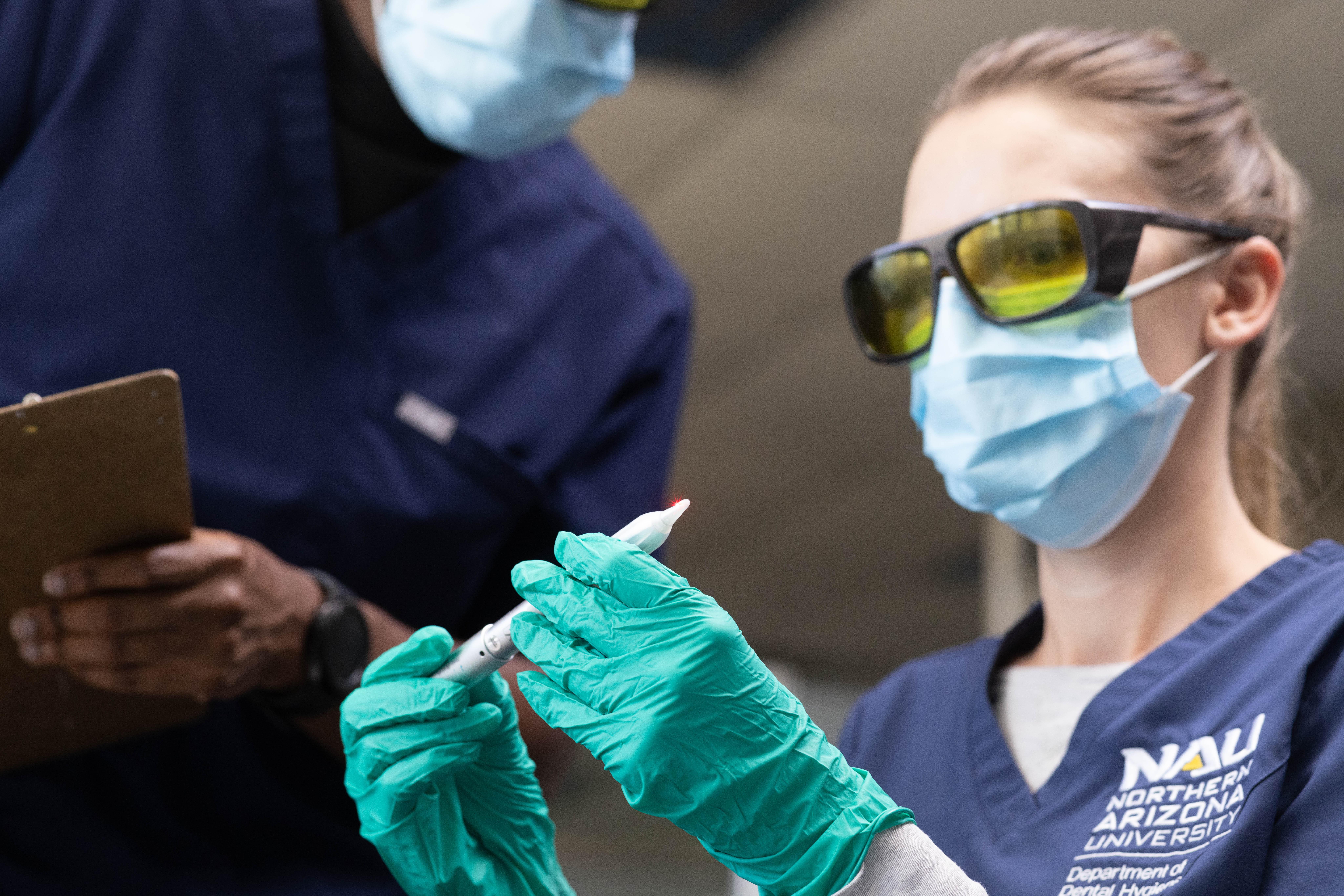 Two medical professionals wearing masks and gloves, one holding a tablet device, the other preparing a syringe. Uniform with medical logo visible. Protective equipment and medical setting.