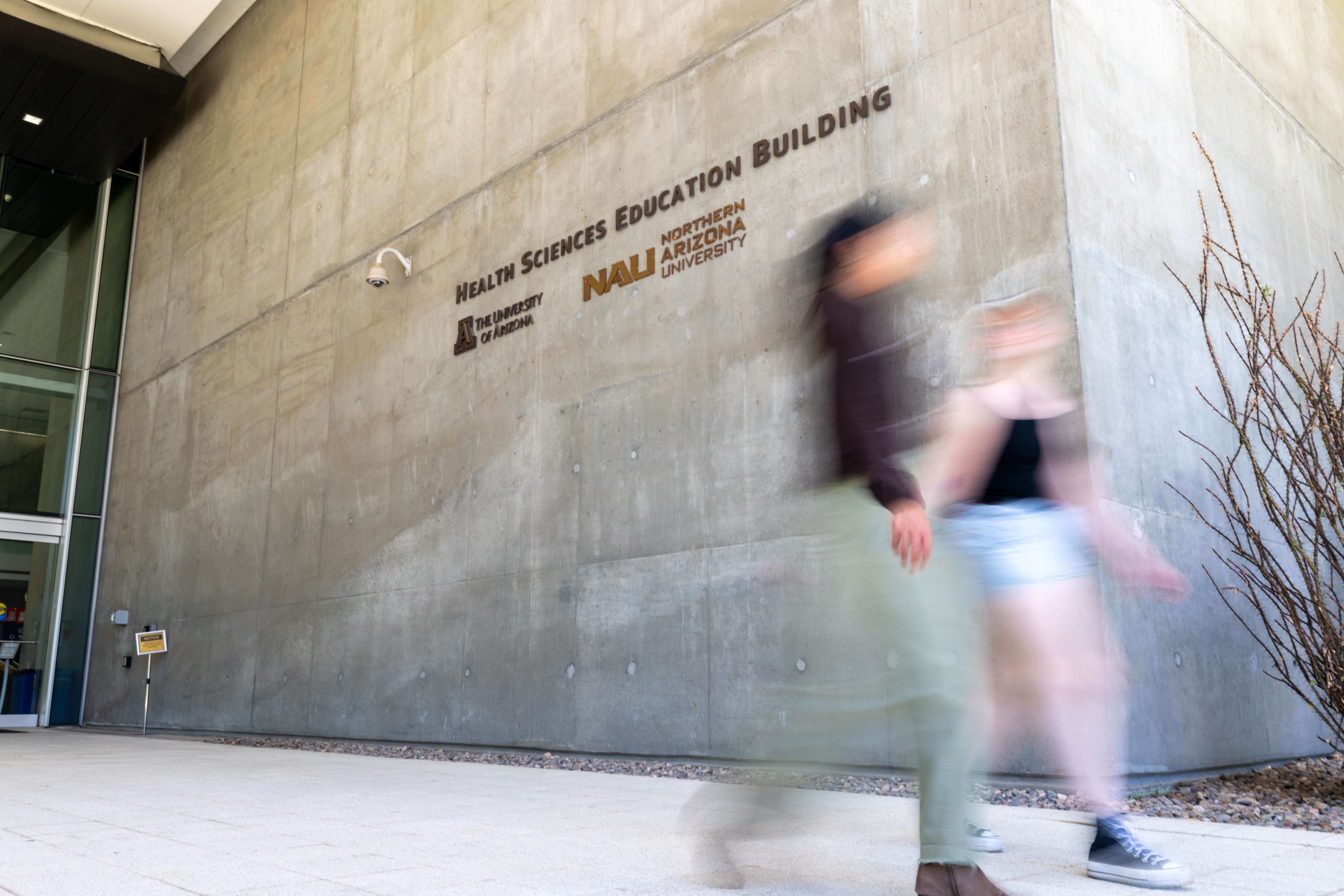 Blurred figures moving past concrete building with text "Health Sciences Education Building" and "Mall Entrance" on wall.