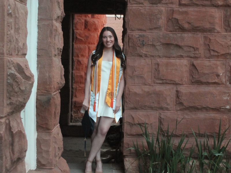 Cidney Roper in graduate regalia and a white dress next to Old Main.
