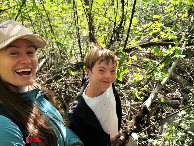 Ella Welsh and young OT client walking on a trail in the woods and smiling at the camera.