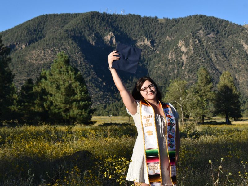 Isabella Guzman posing with graduation regalia in a meadow in front of Flagstaff mountains and blue sky.